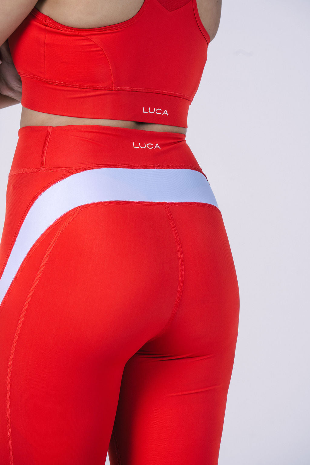 A close-up-shot-from-the-back-of-a-woman-wearing-a-bright-red-athletic-set-showing-the-cycling-bra-and-high-waisted-legging-The- leggings-feature-a-distinctive-wide- white-diagonal-stripe-across-the- rear-The-LUCA-brand-logo-is-visible-on-both-the-bra-band-and- the-top-of-the-leggings-Focus-on- red-workout-leggings-high-waisted- gym-tights-and-women's-matching- athletic-sets