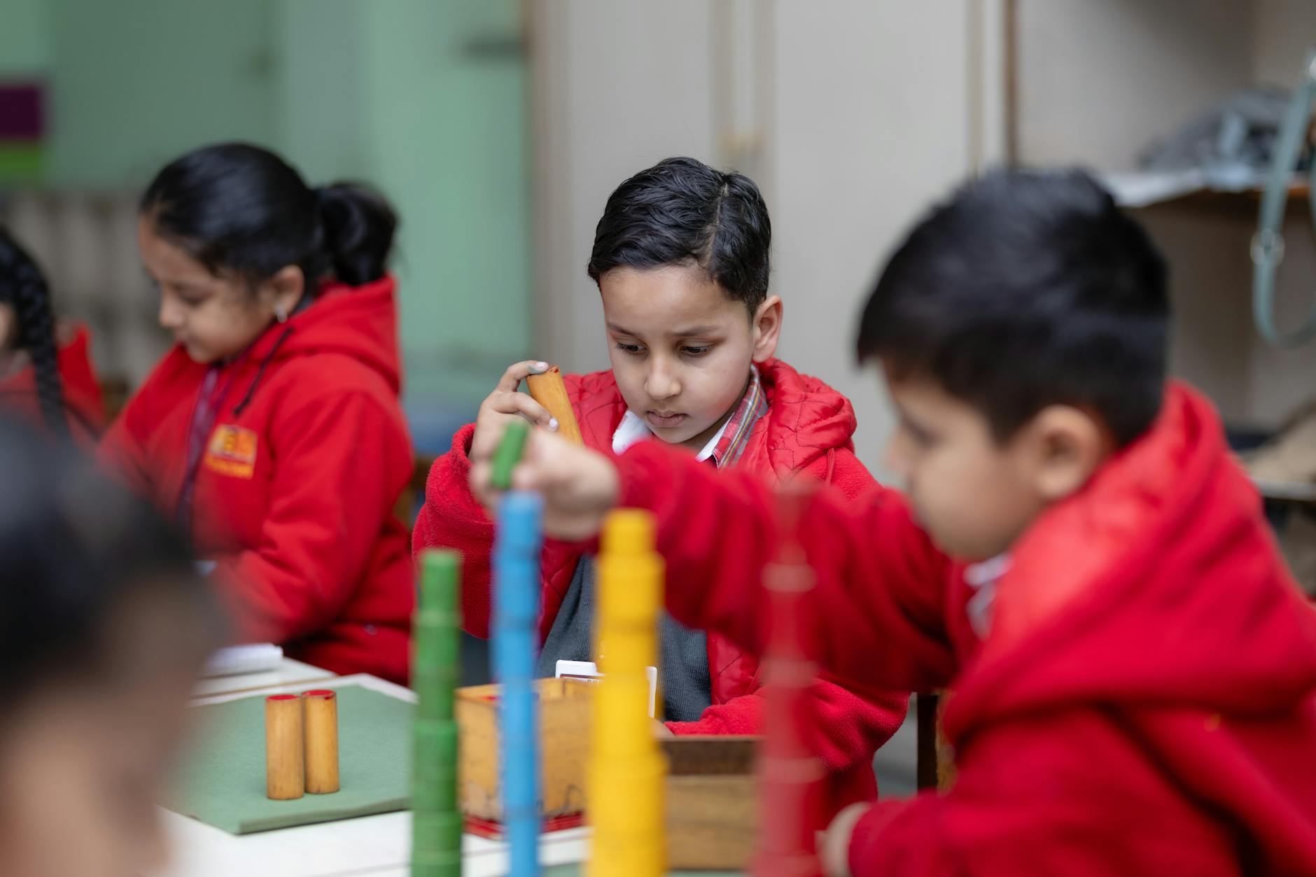 A smiling young girl in a yellow shirt solves a colorful math puzzle at a bright desk in a primary classroom.