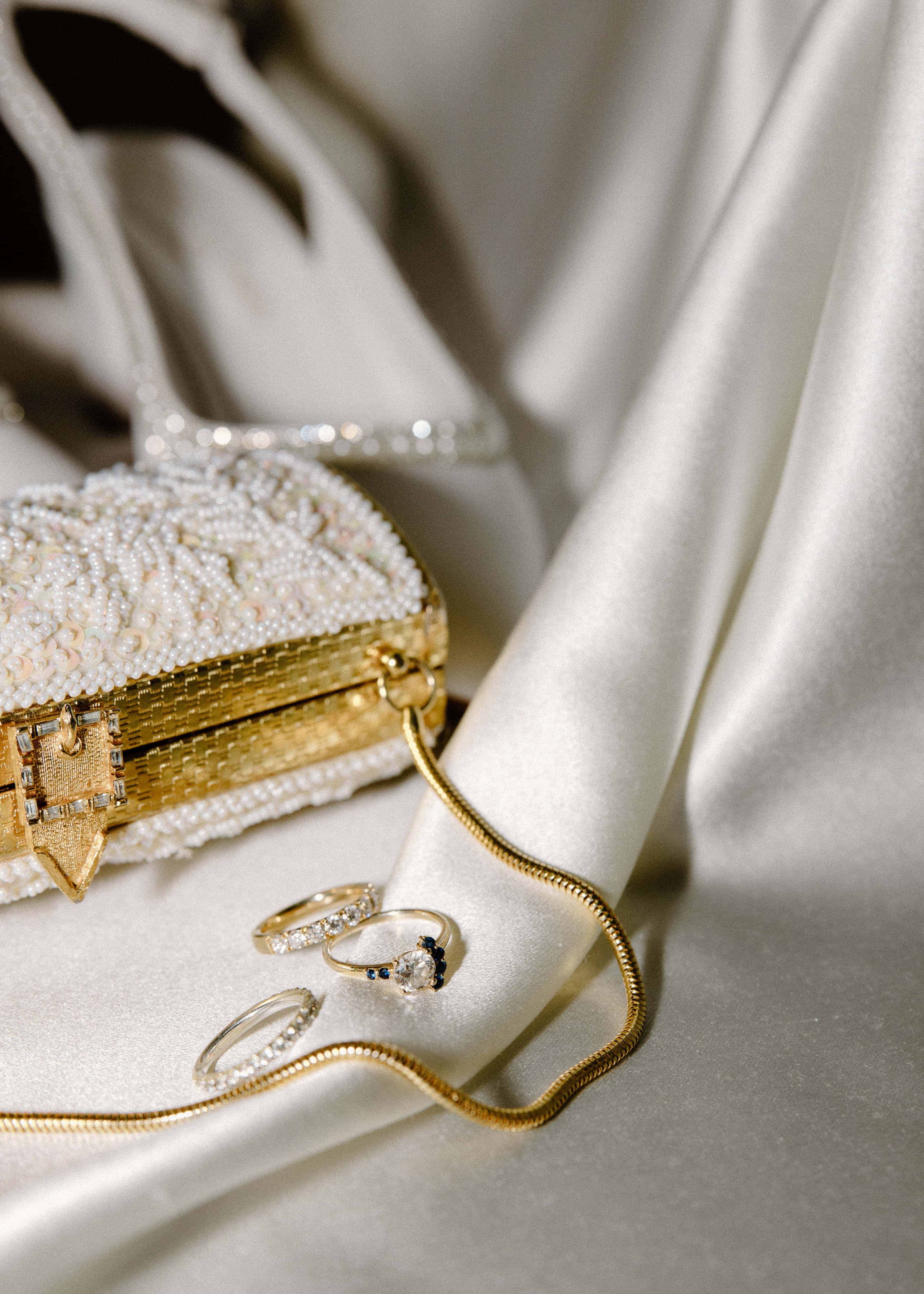 A close-up shot of a ring box on a white silk cloth. The box is open, revealing two gold wedding bands inside.