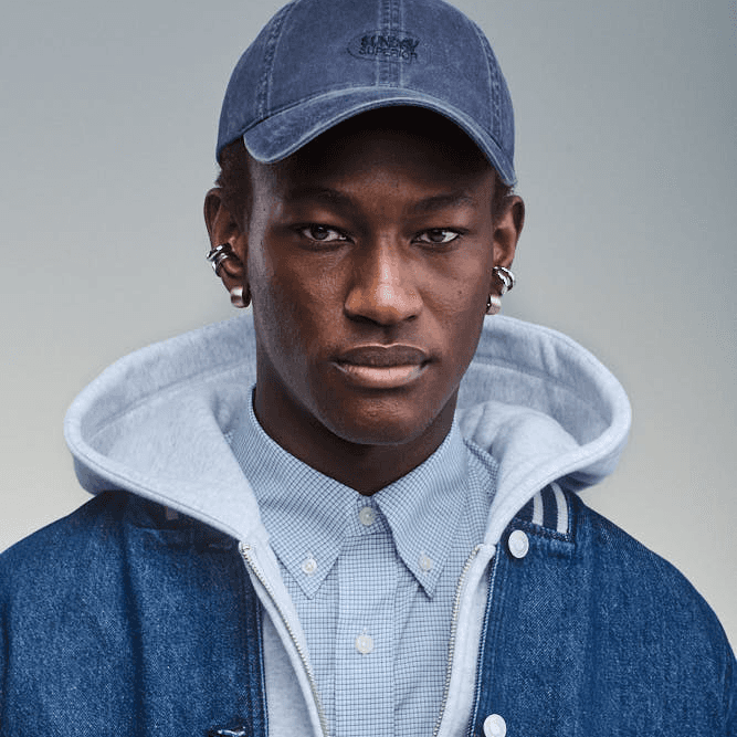 Portrait of a man wearing a light grey Sunday Superior Zip Hoodie, denim jacket and a washed blue Sunday Cap