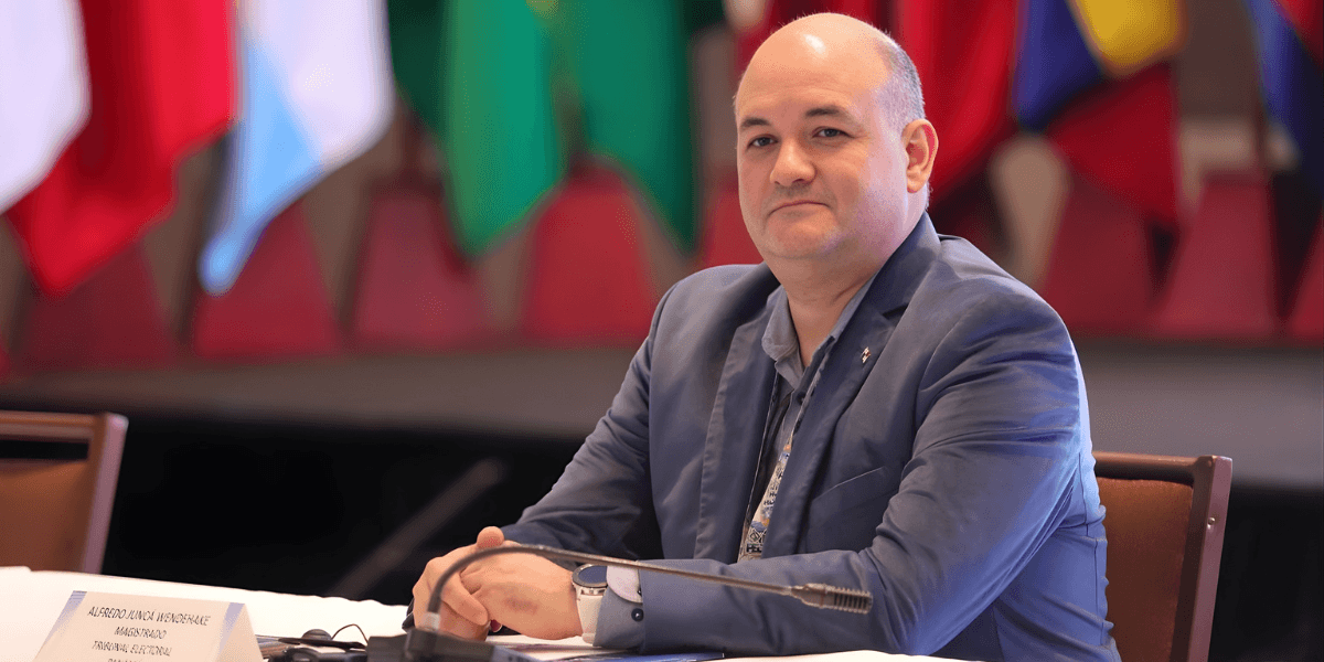 Dr. Alfredo Juncá seated at a table with flags behind him, representing his role in advancing democratic governance in Latin America.