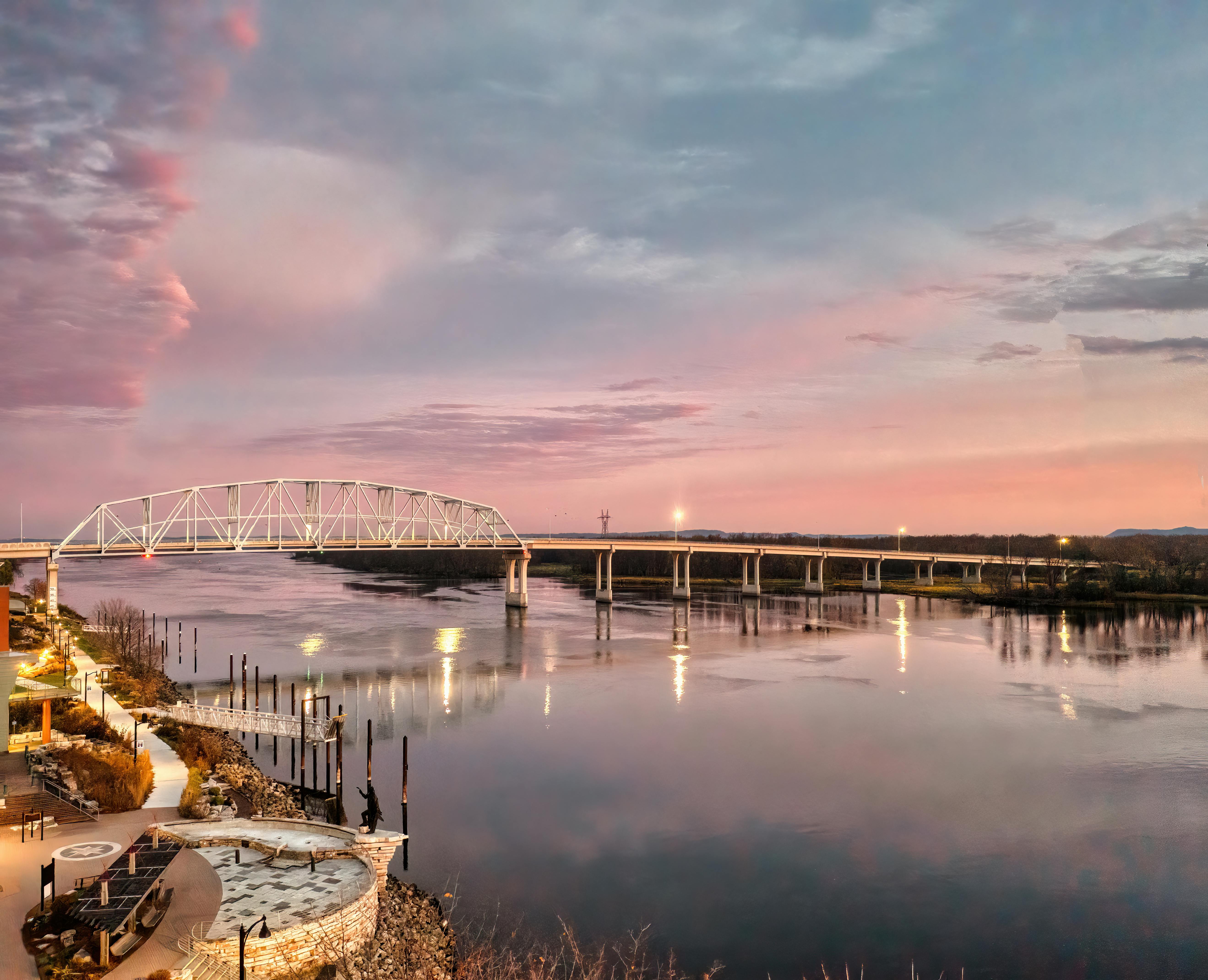 Truss bridge over a wide river at sunset, with illuminated walkway and reflections.