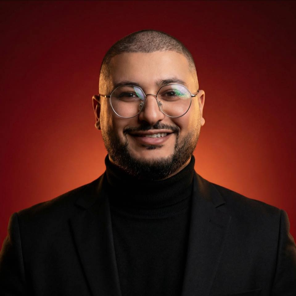 A young man with light brown hair is wearing a black suit jacket and white shirt, standing in a softly lit office setting, conveying professionalism.