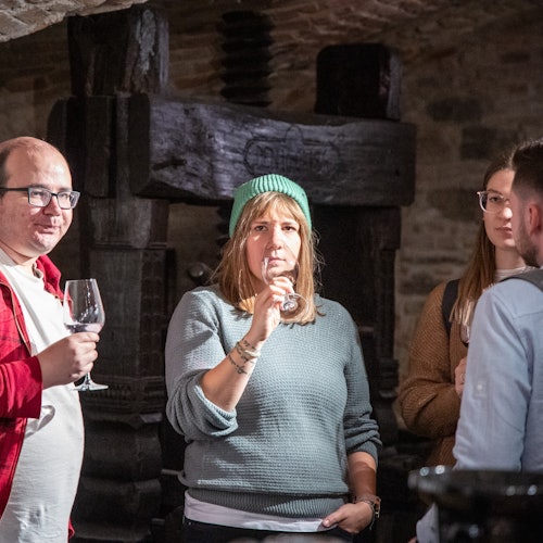 Cuatro personas están de pie dentro de una sala con paredes de piedra, cada una sosteniendo una copa de vino, con una persona bebiendo y una vieja prensa de vino al fondo.