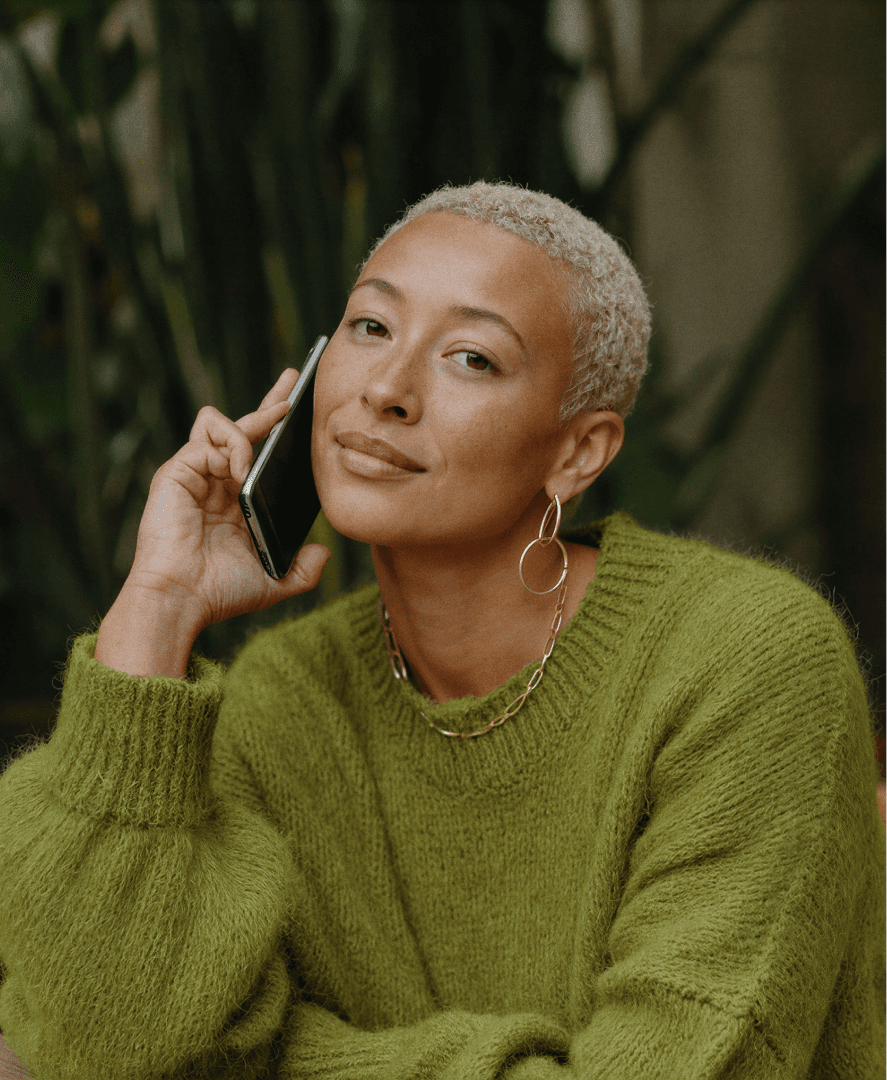 Mulher sentada em cadeira de madeira, com celular no ouvido e expressão serena, com folhas e vasos ao fundo de uma clínica automatizada pela Tivita