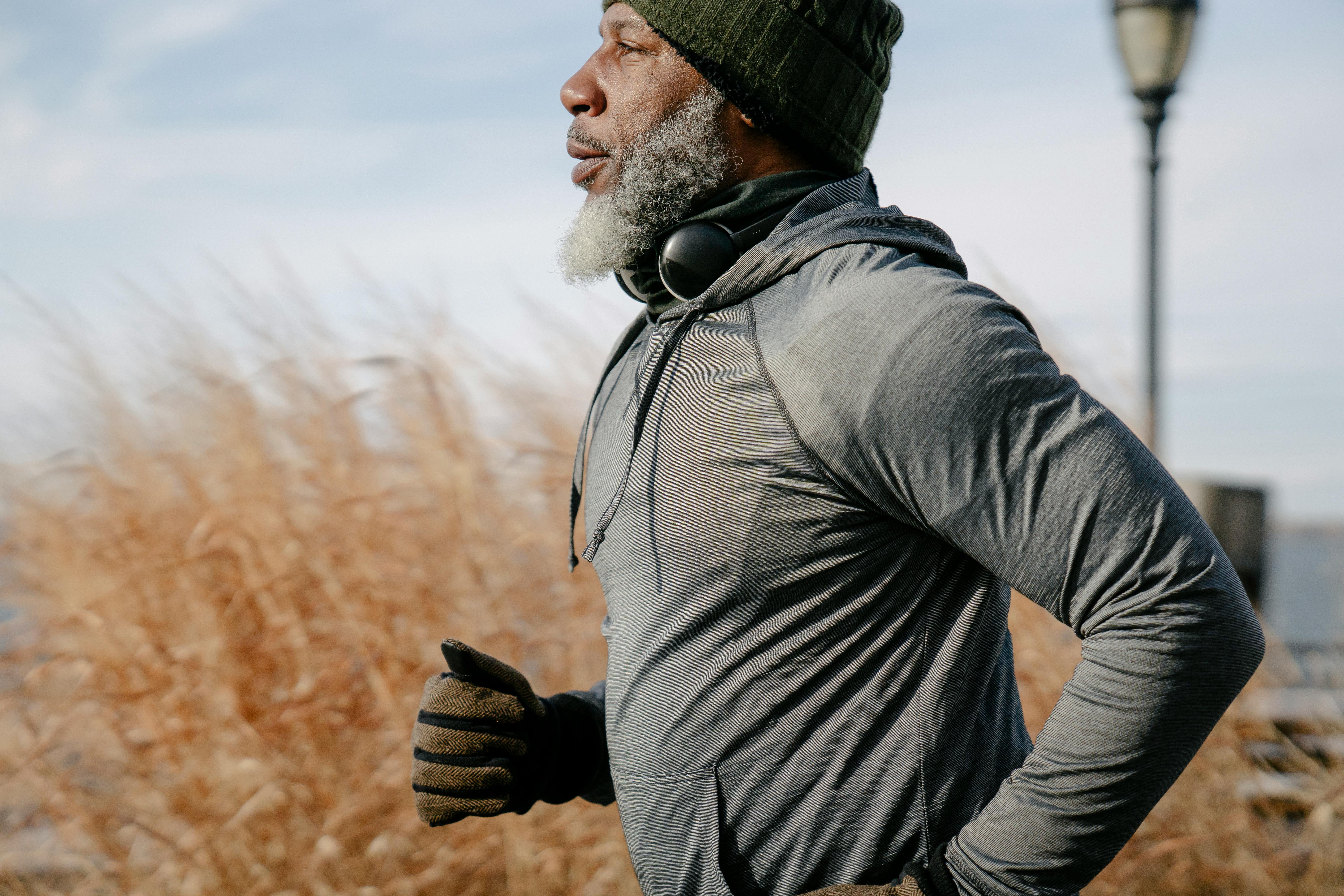 Older man with gray beard, beanie, and headphones jogging.