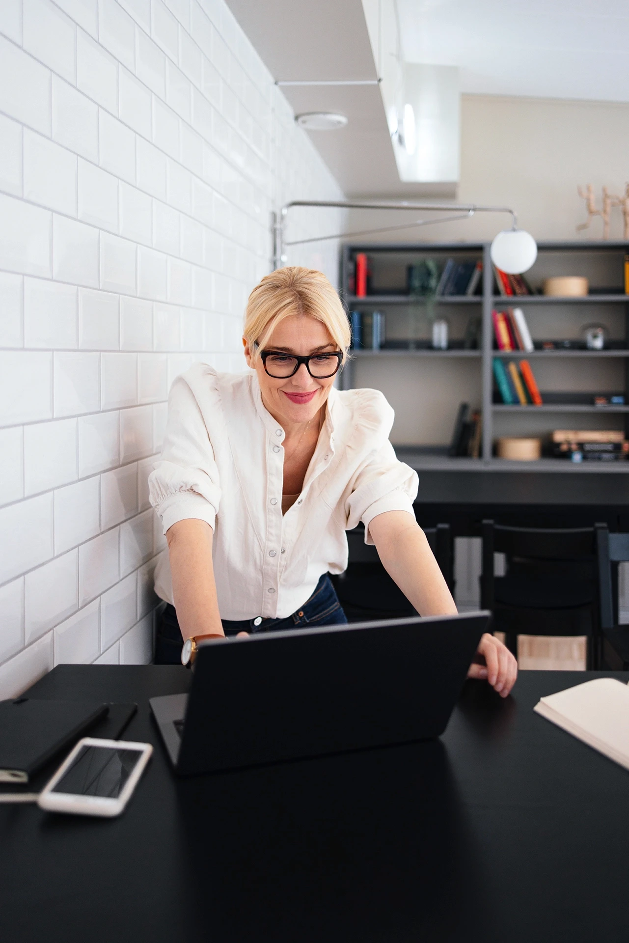 Individual working on a laptop at a table in a home office with shelves and books in the background