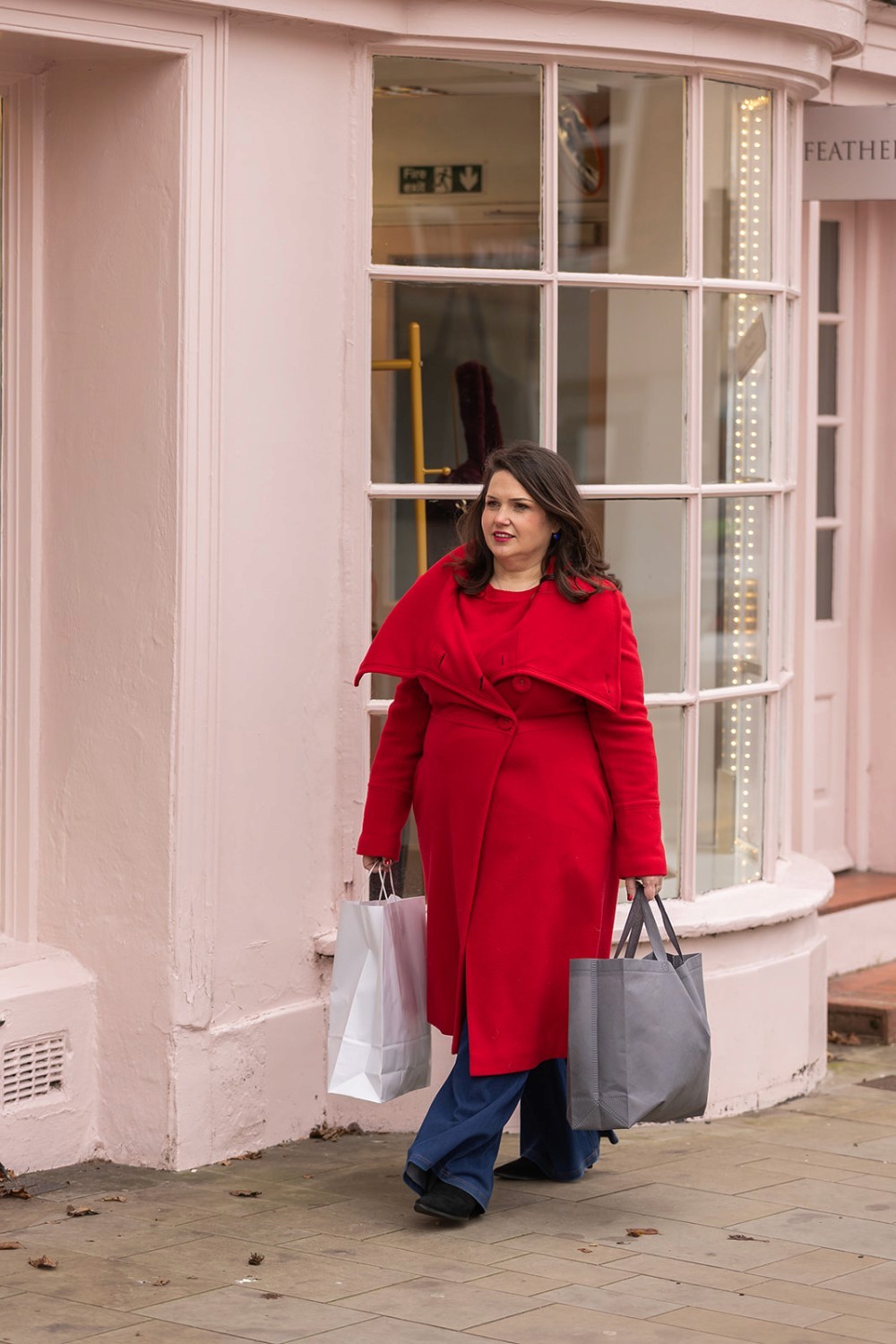 Lucy in a cool toned red coat, walking past Feather and Sole with shopping