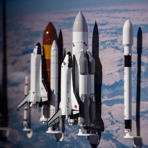 Model rockets and space shuttles mounted on a wall, casting shadows against a backdrop of a blue and white sky.