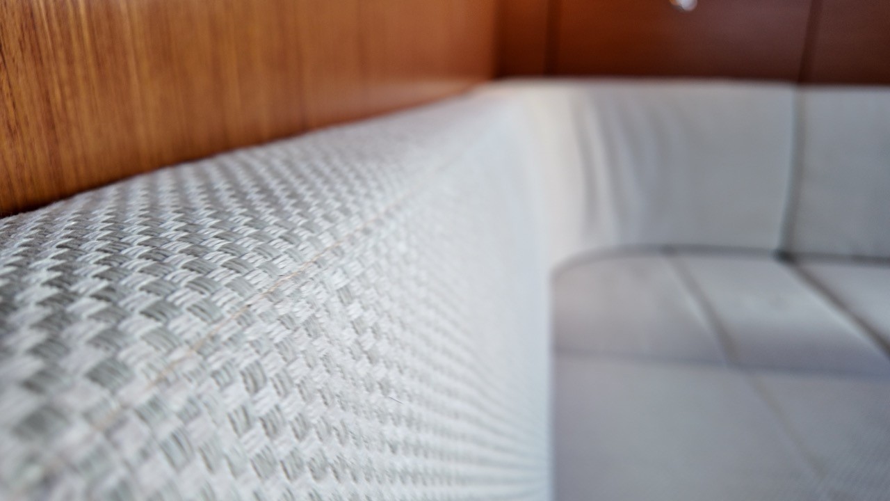 Close-up detail of a self-tailing cockpit winch on the Beneteau Clipper 361 sailing yacht, with a braided red-and-white halyard wrapped around the drum, stainless steel self-tailer jaw, teak cockpit sole and side deck visible in the background over sparkling harbour water