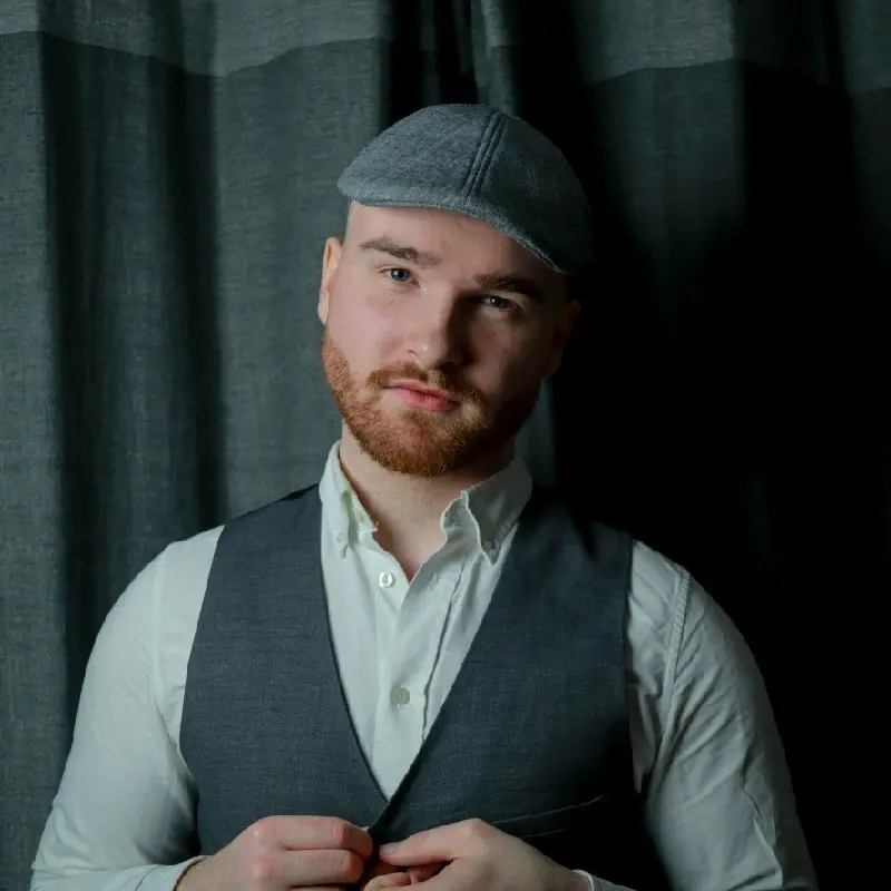 Man with a reddish beard and gray flat cap wears a vest over a white shirt, standing against dark curtains