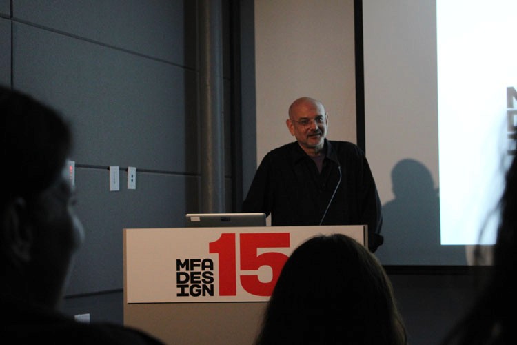 A photo of a man giving a lecture while sitting at a stand with text MFA DESIGN 15 on it.