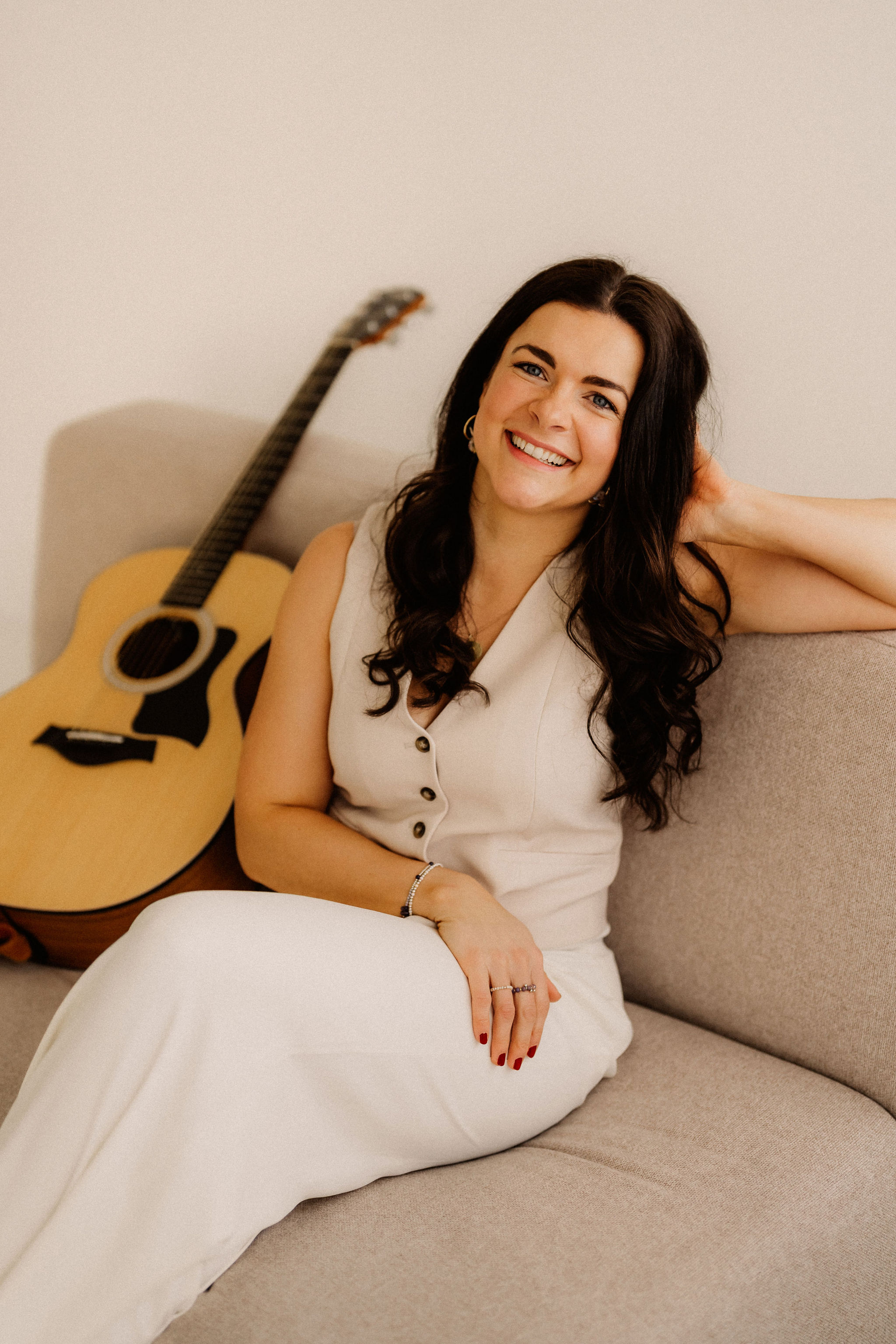 Maria Wiethaler freie Rednerin und Sängerin für Hochzeiten, Taufen, Trauerfeiern und Events aus München und Landshuter Umgebung. Eine fröhliche Musikerin sitzt mit einer Akustikgitarre auf dem Schoß im Freien. Sie trägt ein rotes Kleid mit weißem Muster und hat eine Sonnenbrille auf dem Kopf. Im Hintergrund stehen ein Baum mit weißem Stoff als Dekoration sowie eine grüne Landschaft. Sie lächelt strahlend in die Kamera, während sie sich auf ihren Auftritt vorbereitet.