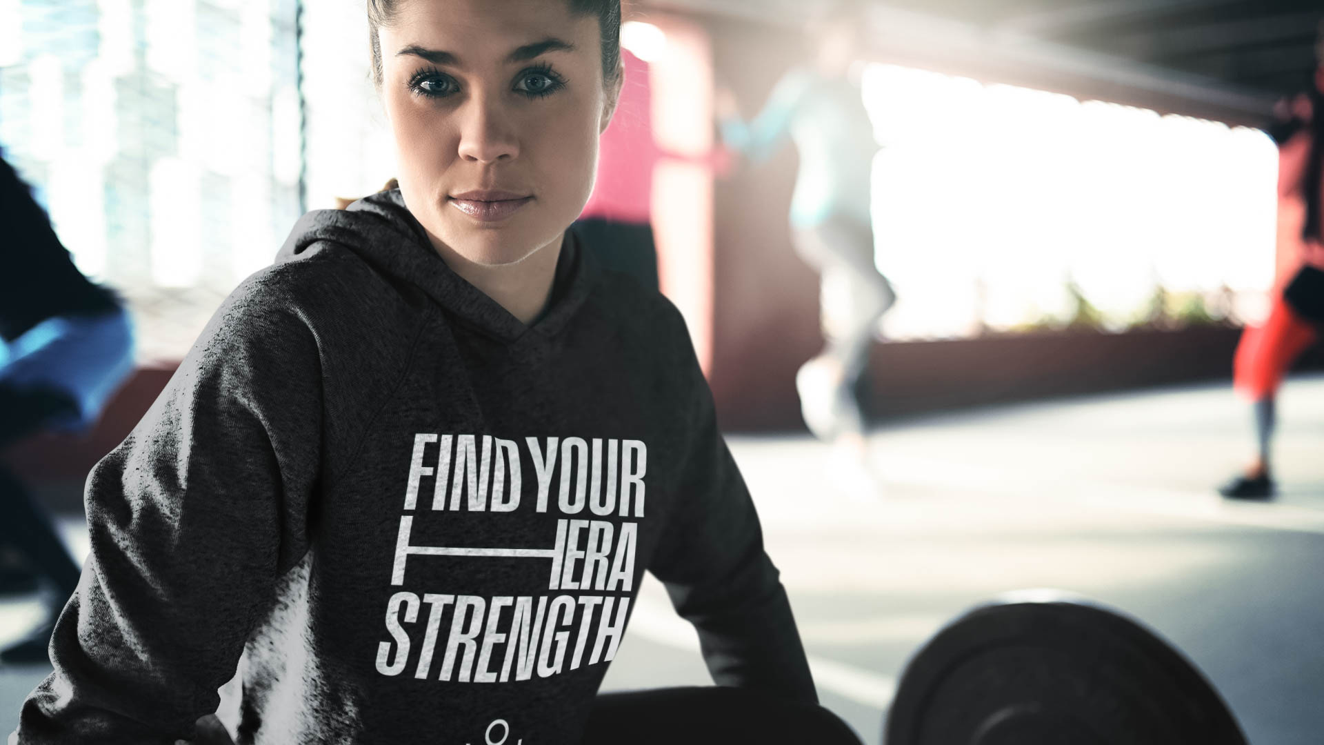A woman with short hair and dark eyes wears a dark gray hoodie with the white text "FIND YOUR HERA STRENGTH" printed on it. She is in a gym setting, with blurred figures of other people in the background. A portion of a weight plate is visible in the foreground. The gym floor is light-colored.