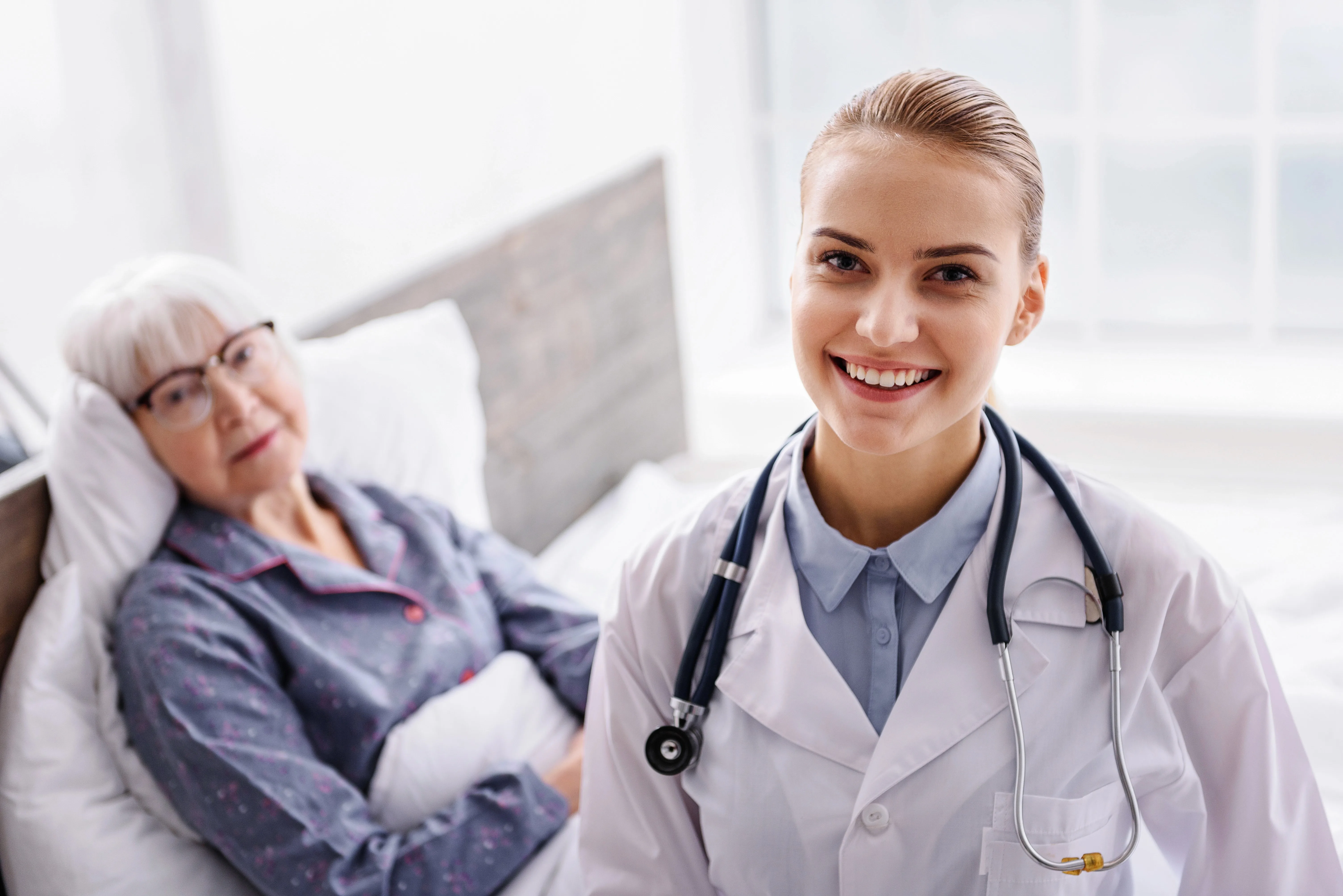 White female physician smiling, wearing white coat with stethoscope around her neck, old female patient with eyeglasses laying in bed]