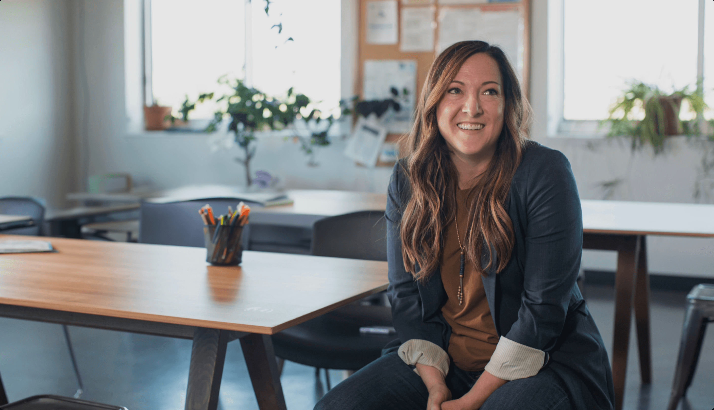 A smiling woman sits at a wooden table in a bright room with plants, holding a coffee cup.