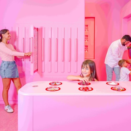 一位女士与墙壁互动，一名女孩在粉色柜台上伸手拿物品，一位男士带着孩子们探索一个粉色房间。
