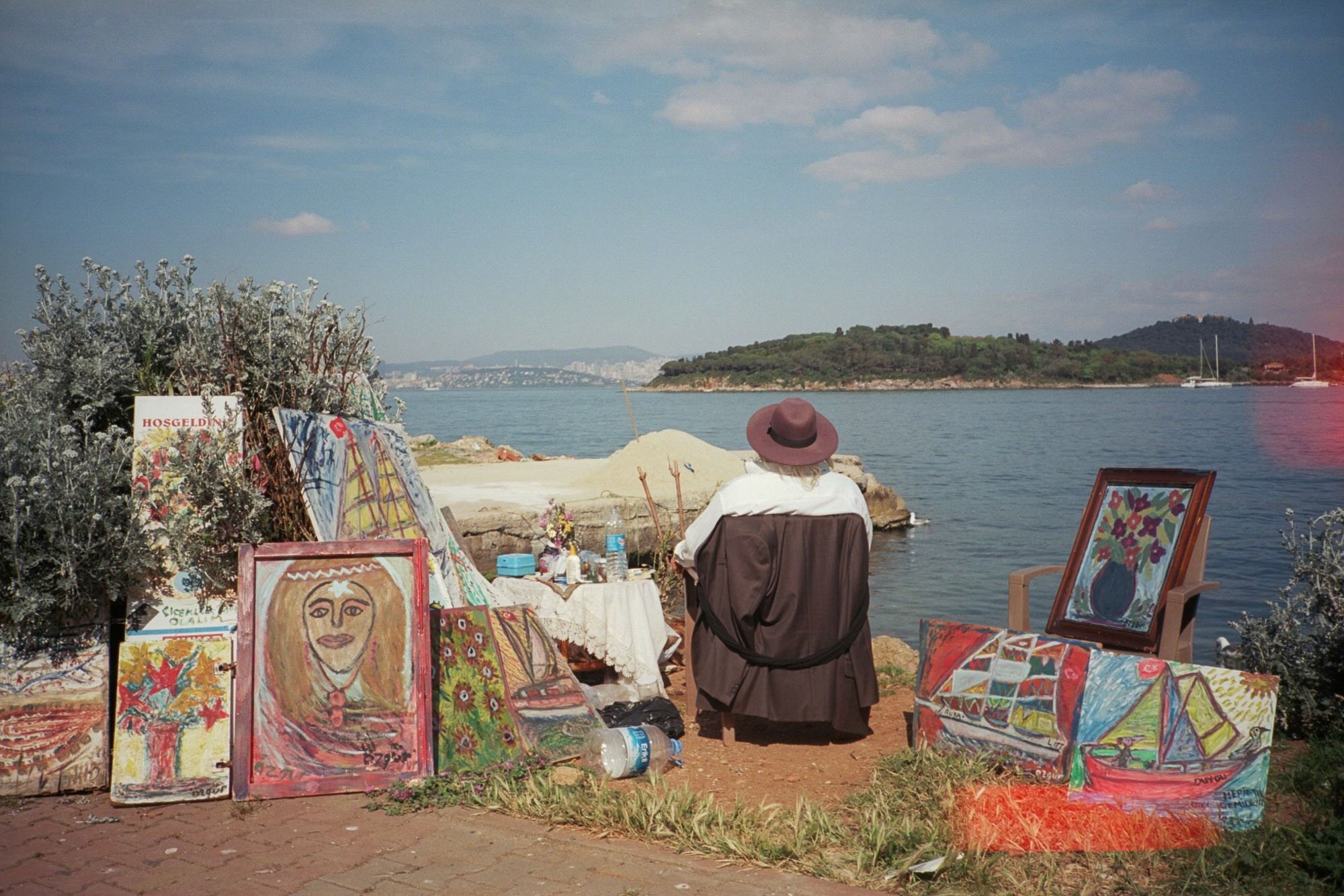 A person wearing a hat sits in front of a collection of colorful paintings displayed along a scenic waterfront with lush greenery and boats in the background.