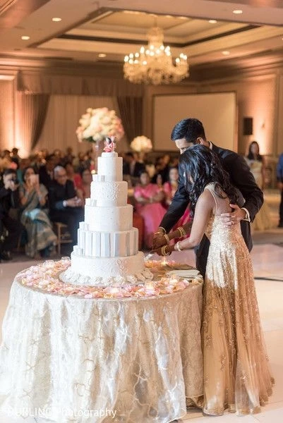 Bride and groom cutting a tall, multi-tiered white wedding cake adorned with flowers, surrounded by guests and soft lighting, capturing a formal and elegant wedding reception moment by Out of The Blues Fine Art Wedding Photography in Hyderabad.