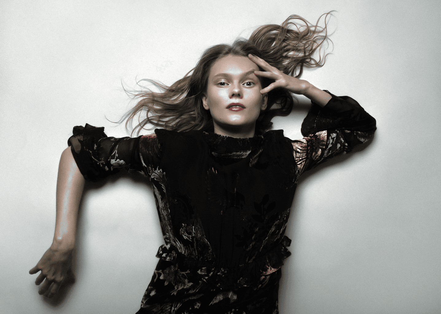 Portrait of Icelandic musician Sunna Margrét lying against a light backdrop in a dark patterned dress, her hair spread out dramatically around her head. Photo by Olivia Schenker