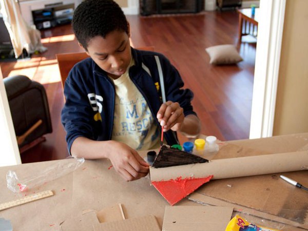A young boy with dark hair is seated at a table covered in cardboard, working on a craft project. He is wearing a dark blue jacket over a cream-colored shirt with the letters “MA RESC” visible. His right hand holds a red triangular piece of cardboard, and his left hand holds a glue stick. Several small containers of colorful glue are on the table near him. The table surface is scattered with cardboard pieces. A silver pen lies on the table near the right edge. The background shows a hardwood floor, a portion of a couch, and a doorway.