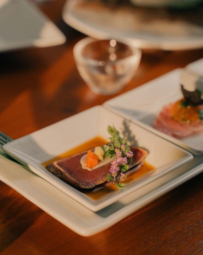 A beautifully plated dish featuring thinly sliced fish garnished with colorful toppings and herbs, served on a rectangular white plate.