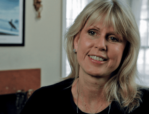 A woman with shoulder-length blonde hair smiles at the camera. She wears a black top and a silver necklace. A framed picture hangs on the wall behind her, and a window is visible to the right.