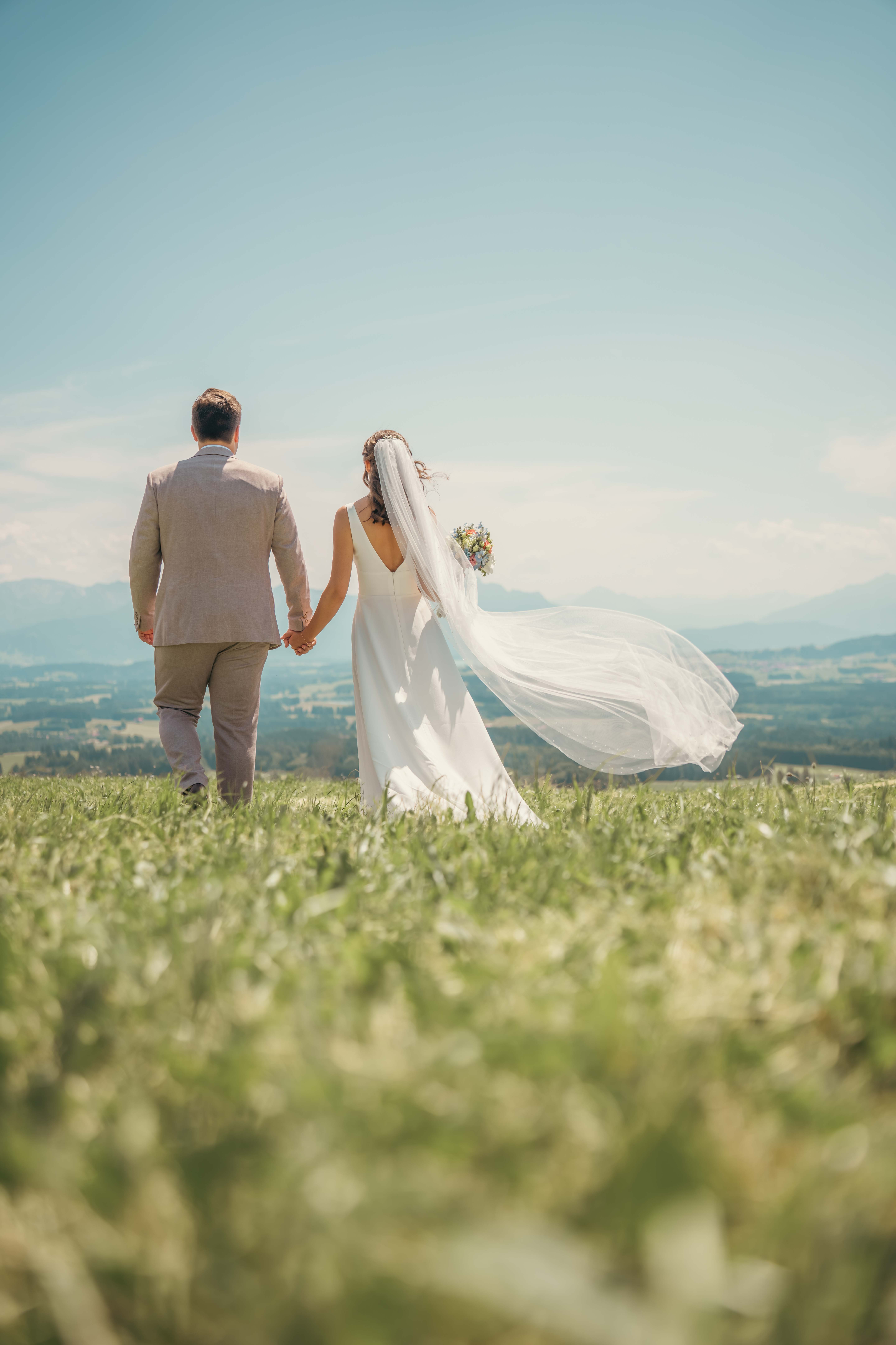 A couple in wedding attire walks hand in hand across a sunlit field.