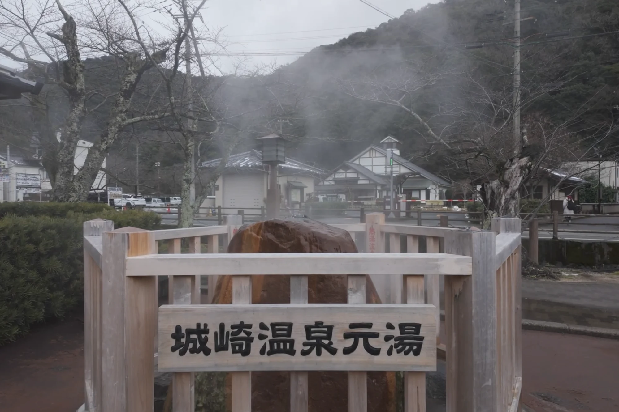 Steam rising from the Motoyu hot spring source beside a Kinosaki street behind a timber rail.