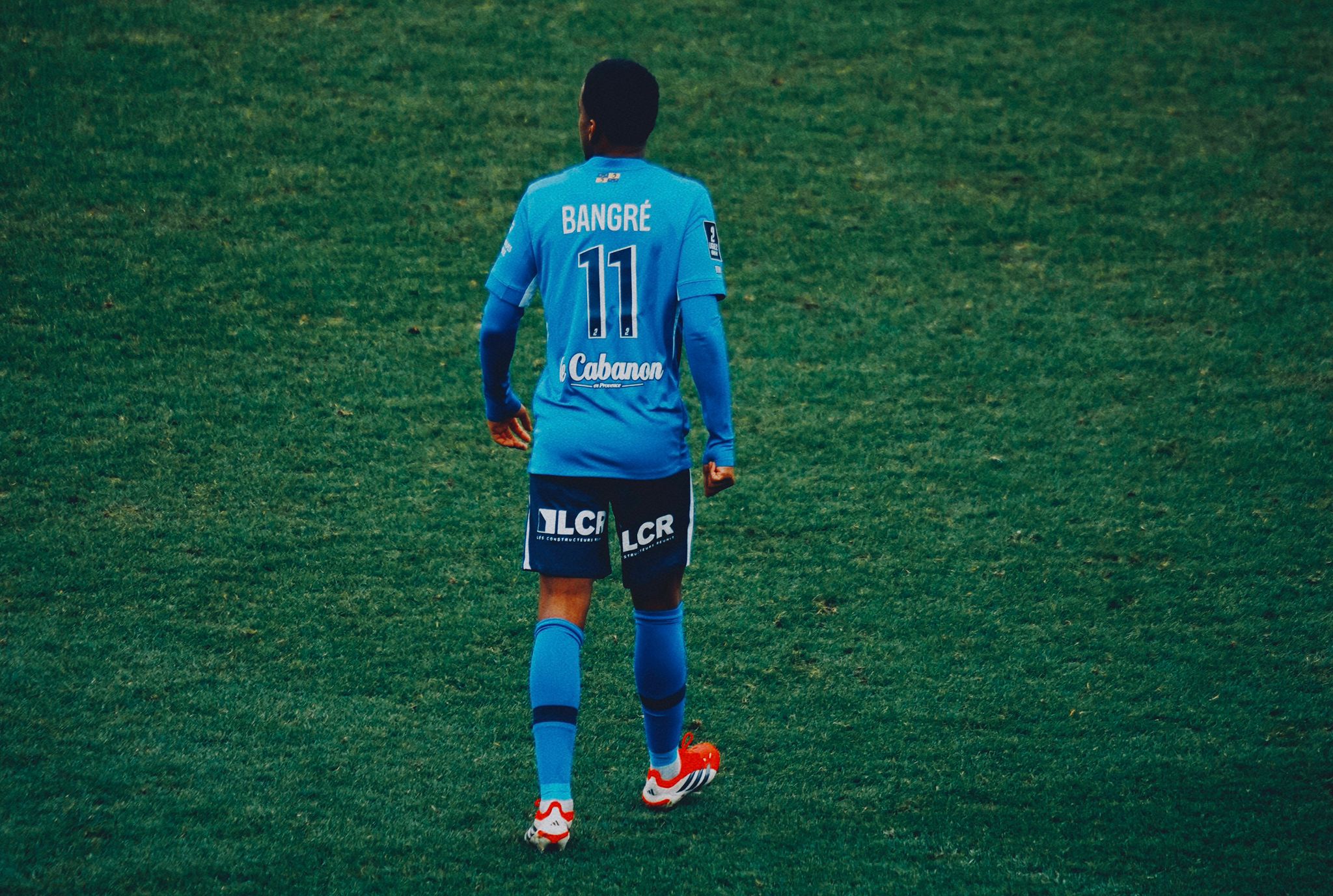 Vue de dos du joueur de football Bangré, portant le maillot bleu numéro 11, marchant sur la pelouse verte lors d'un match.