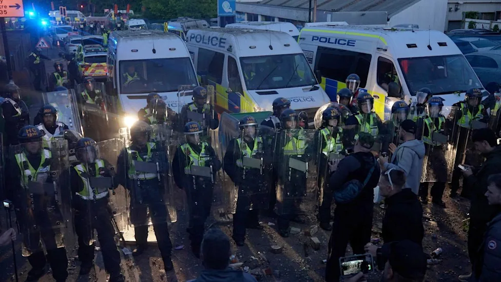 A line of riot police with shields up, opposite pro-Tommy Robinson protesters, a few men wearing casualwear and hats facing the police. Behind the row of riot police are police vans with their headlights on
