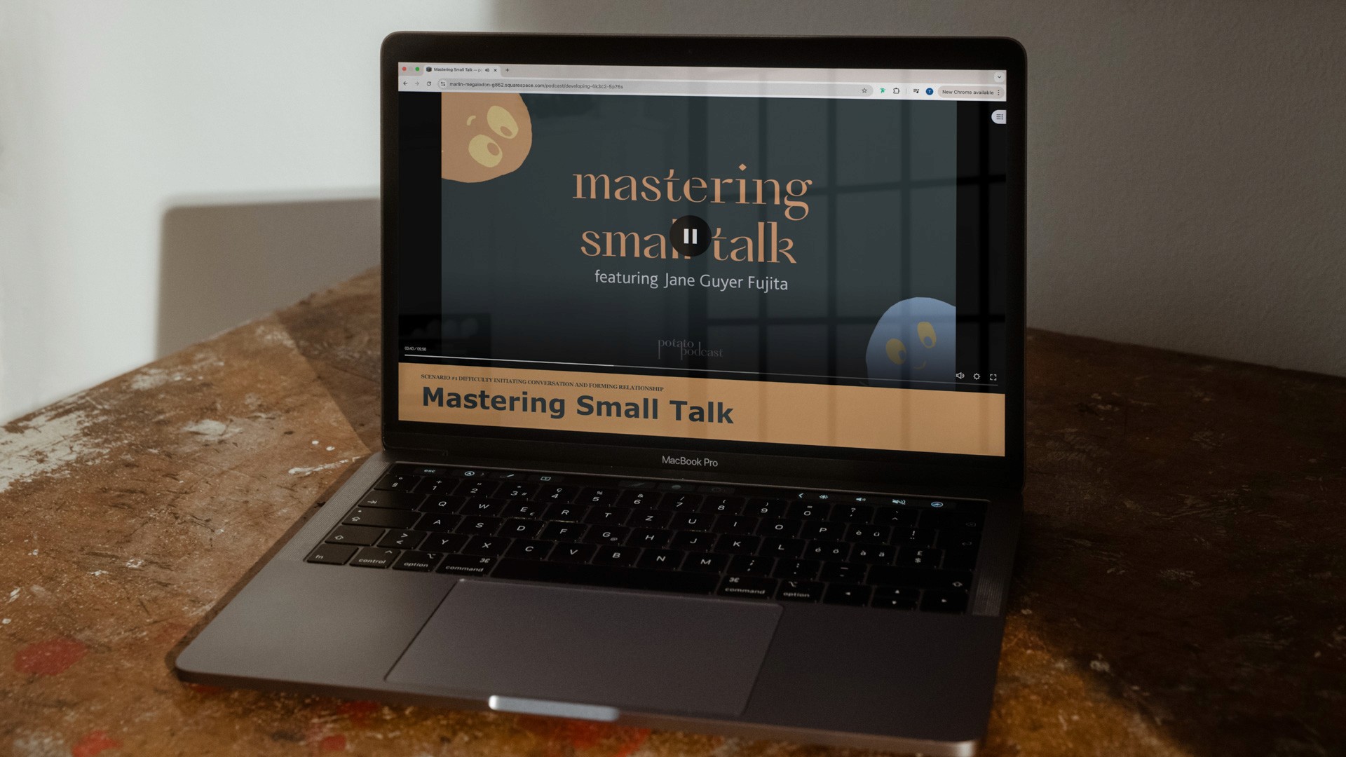 A dark gray MacBook Pro laptop is open on a worn wooden surface, angled slightly to the right. The screen displays the title "Mastering Small Talk" in gold text against a black background. Below the title, smaller text reads "featuring Jane Guyer Fujita." A light tan banner at the bottom of the screen shows "Mastering Small Talk" in bold black text. The laptop's trackpad is visible below the screen. The background includes a white wall and a shadow cast by the laptop.