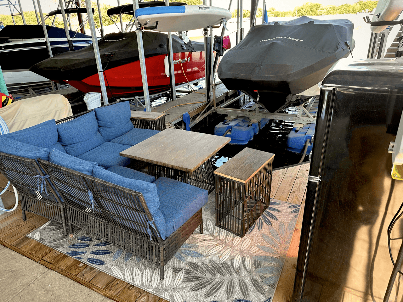 Cozy outdoor seating area on a dock features a wicker sofa set with blue cushions and a wooden coffee table, surrounded by a patterned rug, next to covered speedboats.