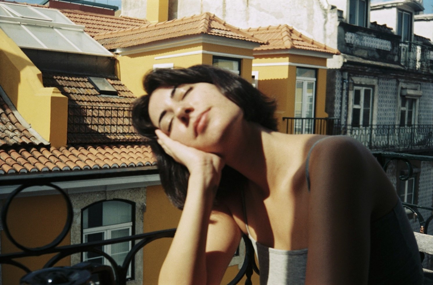 A woman with short dark hair relaxing on an outdoor balcony with a yellow building with tile roofs in the background.
