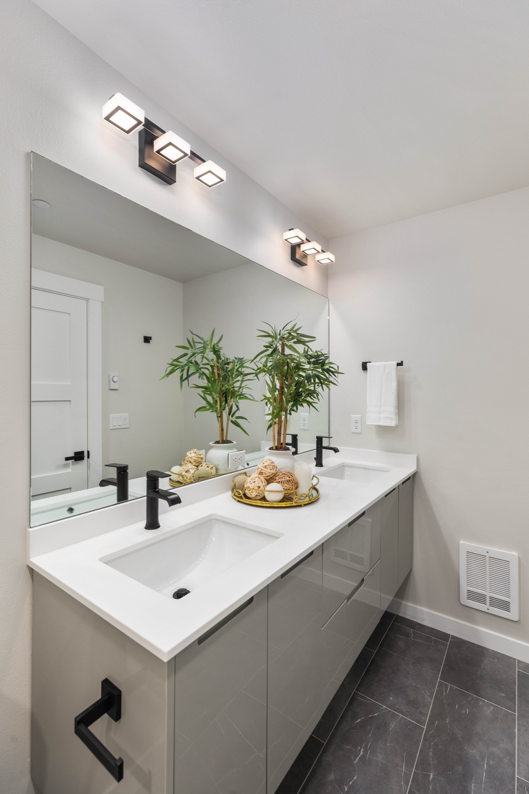 Modern luxury double-sink bathroom vanity with white undermount sinks, grey cabinetry, black fixtures, oversized mirror, and fresh greenery accent