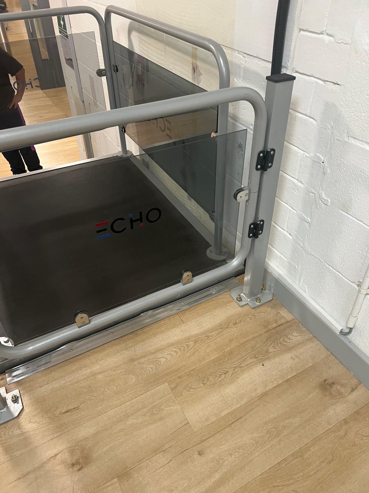 Echo platform step lift with glass side panels and grey steel frame installed in a commercial interior — open platform design for wheelchair access