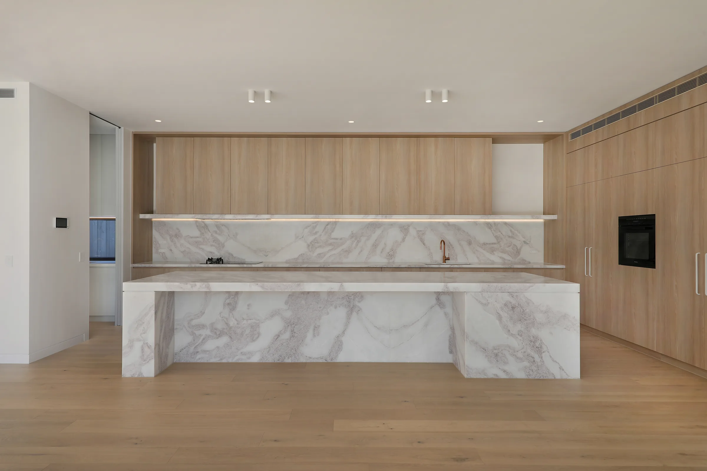 Kitchen viewed head-on with a white marble waterfall island, oak cabinetry, a full-height marble splashback and copper tap over oak floors