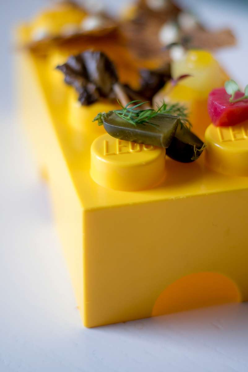 A yellow brick topped with small plastic models of a green leaf, a red berry, and brown dried leaves.