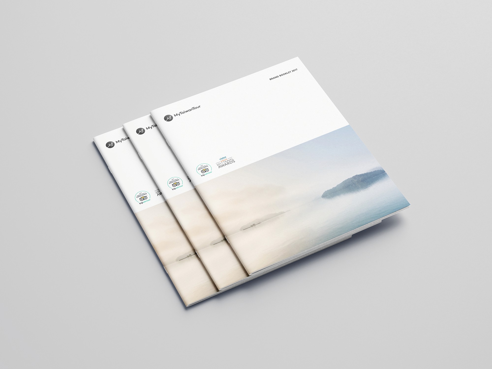 這張圖像展示了 MyTaiwanTour 的品牌手冊,手冊封面設計簡潔,左上角有 MyTaiwanTour 的標誌,底部有一張風景照片,展示了一片寧靜的湖泊和遠山。手冊上還印有一些獎項標誌。