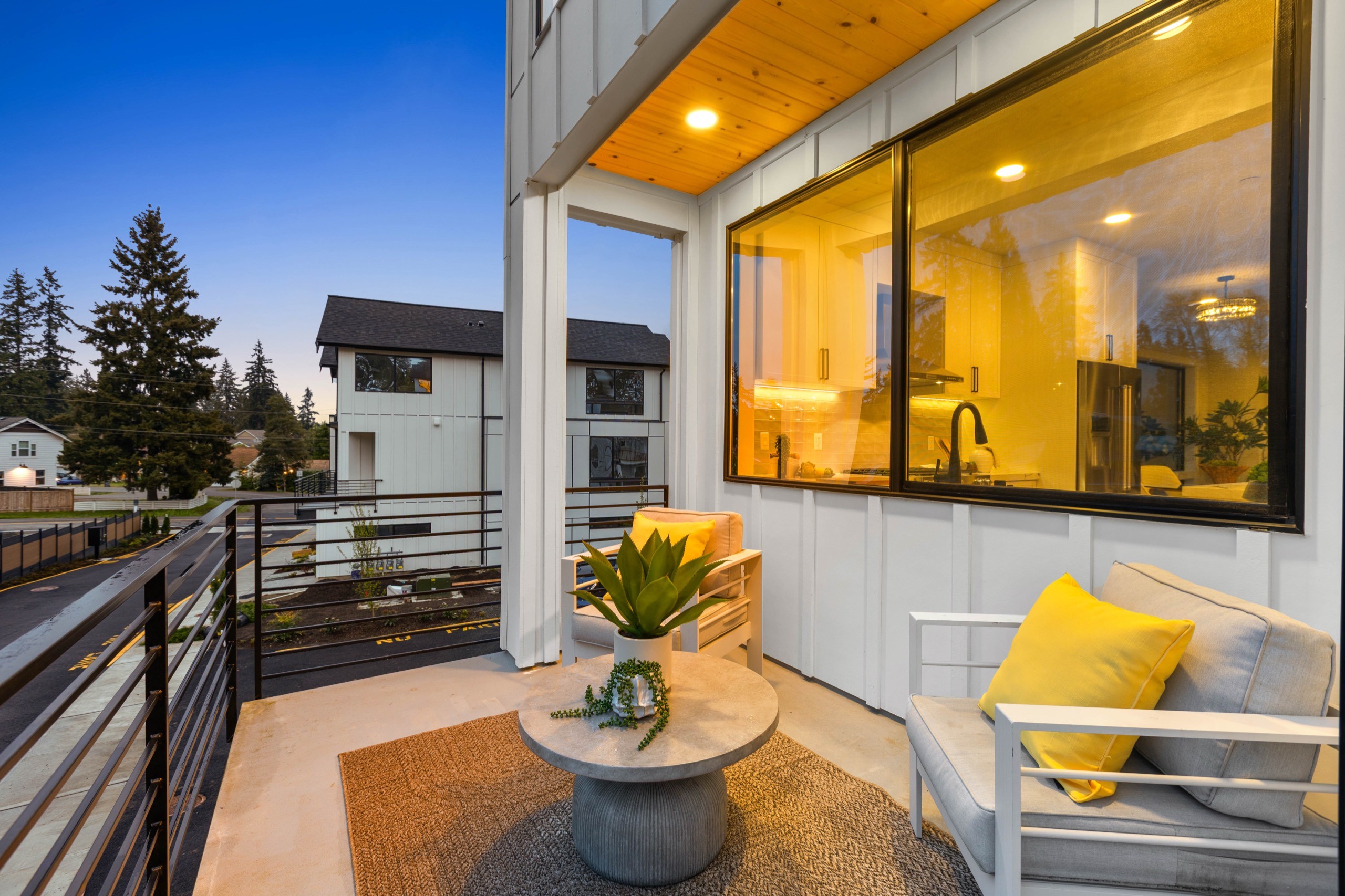 Evening view of a luxury townhome balcony with white lounge chairs, yellow accent pillows, round concrete side table, and warm interior kitchen visible through floor-to-ceiling glass doors