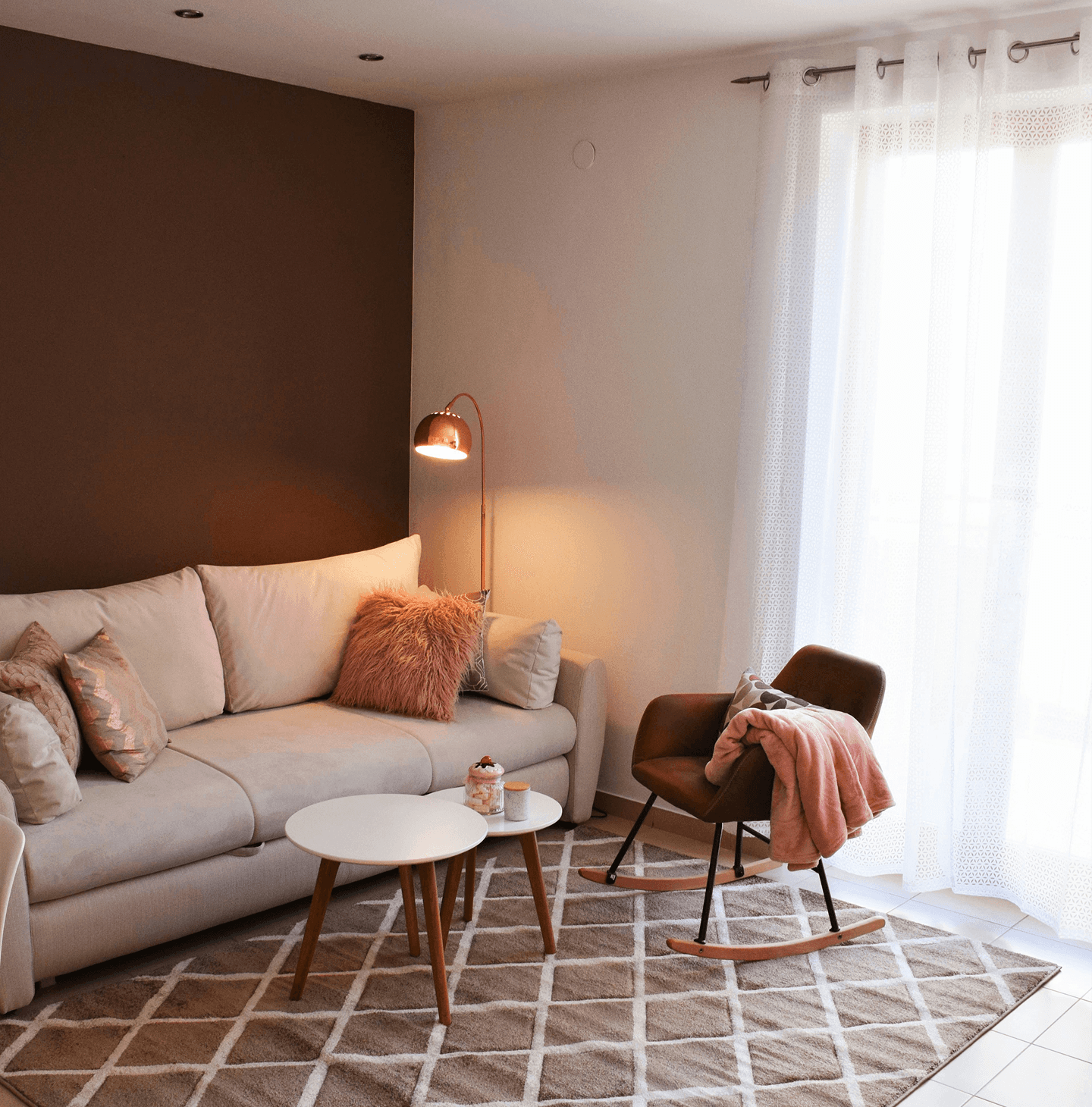 A cozy living room featuring a beige sofa with plush pillows and a furry throw, a modern rocking chair draped with a soft pink blanket, illuminated by warm light from a stylish floor lamp, set against a contrast of dark brown and white walls, with sheer curtains letting in natural light over a geometric patterned area rug.