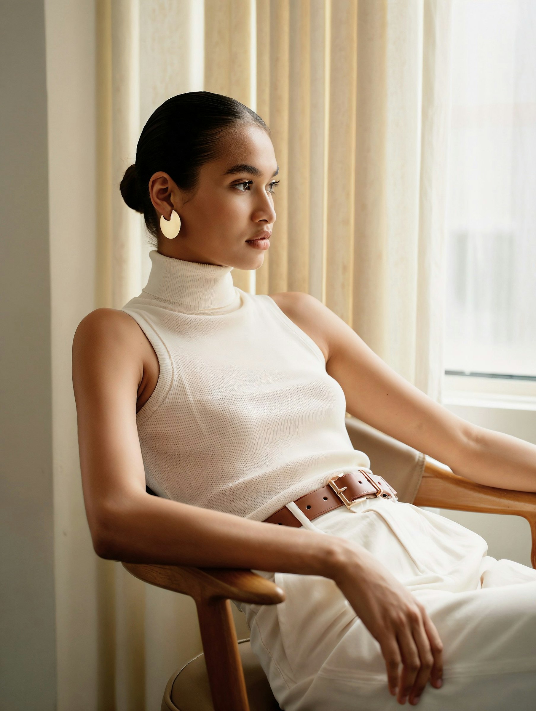 A woman in a minimalist white outfit with gold earrings sits by a window, exuding modern elegance in soft natural light.