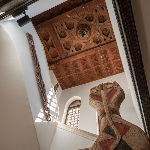 Domed wooden ceiling with intricate designs, large sculpted figure, narrow arched window, and decorative textile hanging on a wall.