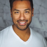 Smiling man with short dark hair and light stubble wearing a white v-neck t-shirt against a gray textured background.
