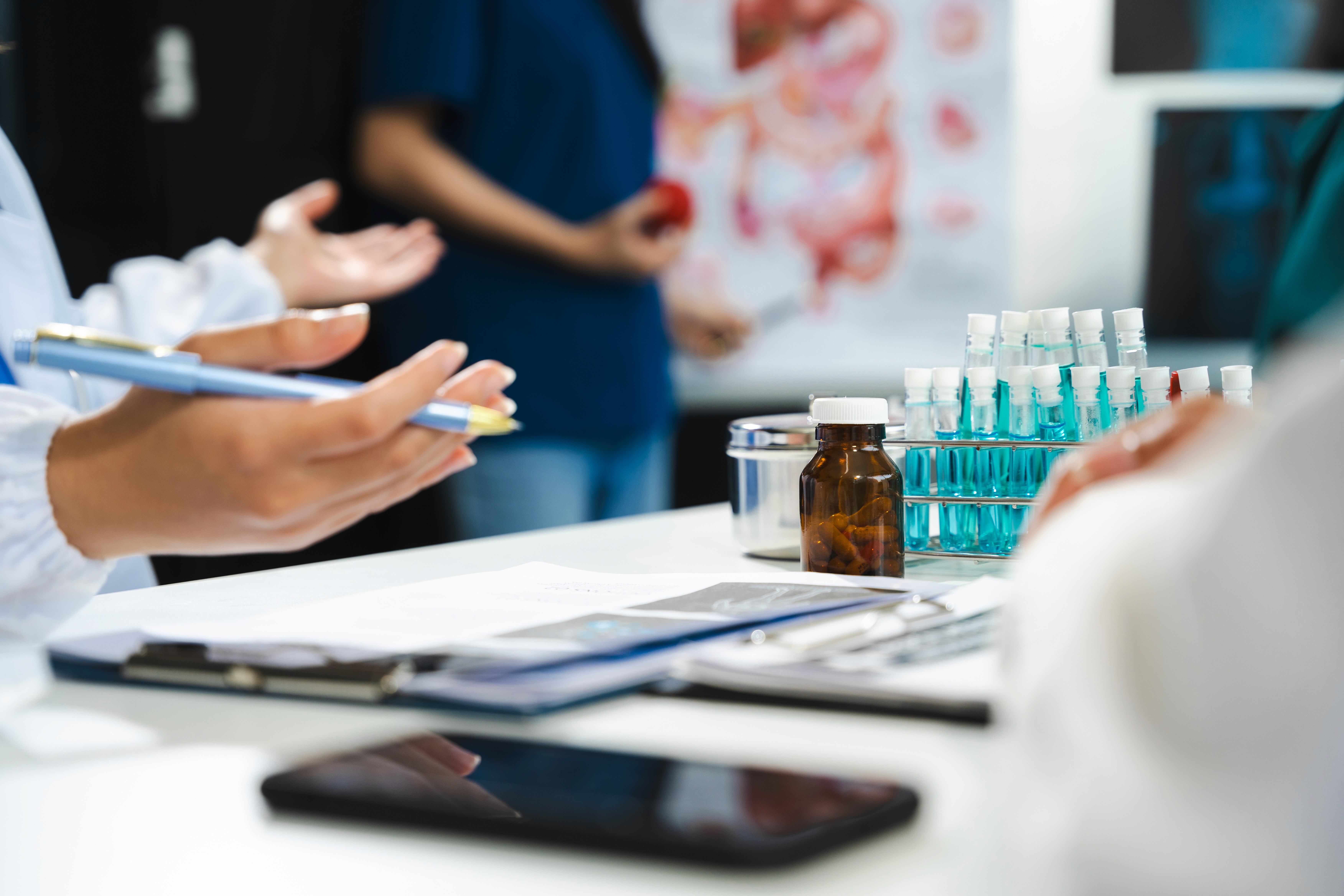 Profesionales sanitarios en una consulta médica con tubos de ensayo y muestras en laboratorio, tomando notas frente a un póster anatómico.