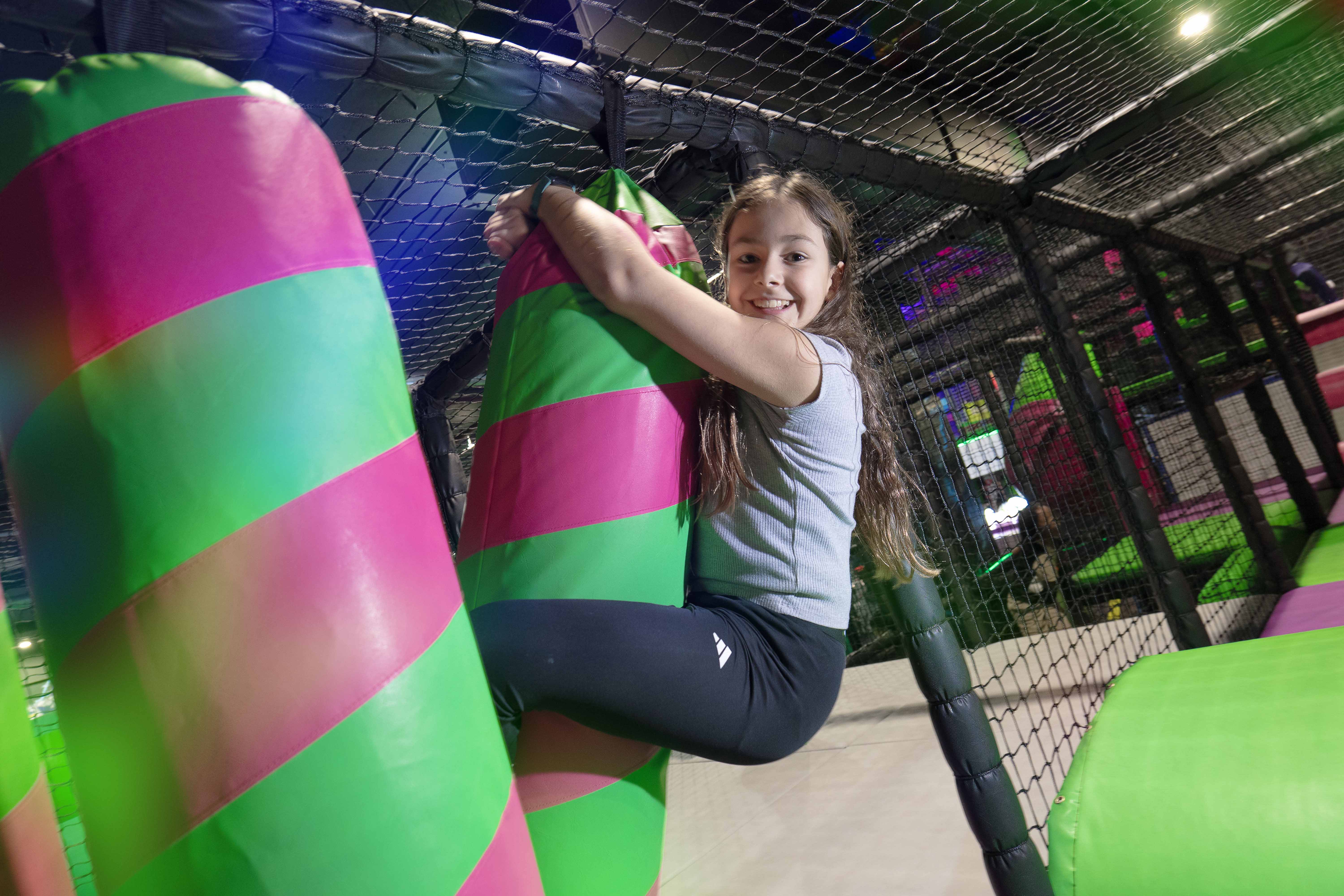A young girl with long hair is joyfully hugging a large, colorful, pink and green striped inflatable column in an indoor play area, surrounded by safety nets and bright, playful lighting.