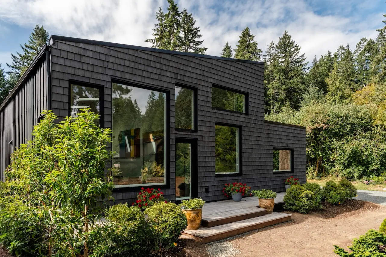 Modern two-story home with large glass windows, warmly lit interiors, and a balcony. Surrounded by trees and a manicured lawn at dusk. Cozy ambiance.