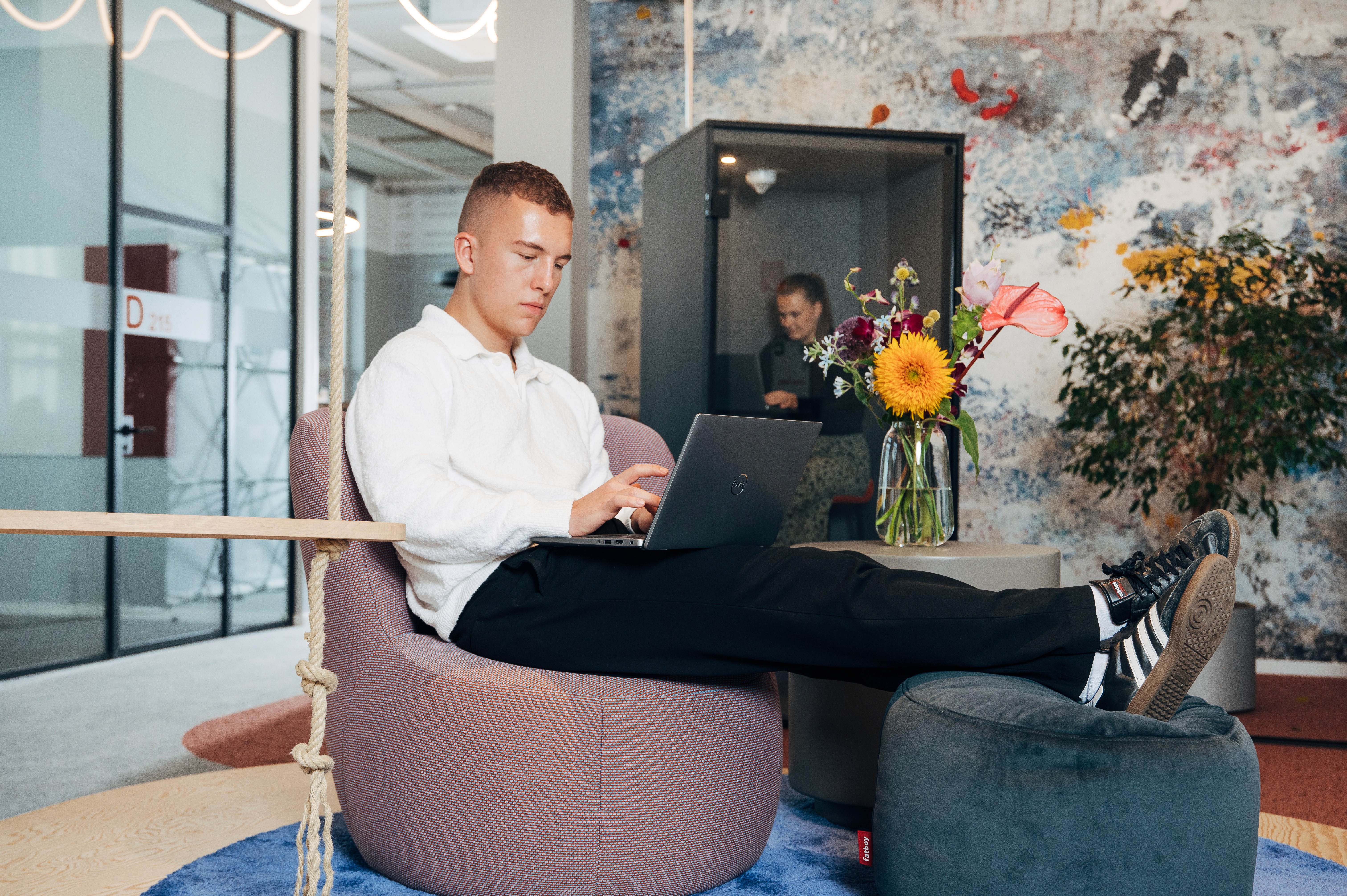 A person sitting in a modern office space, relaxed with feet up, using a laptop surrounded by plants.