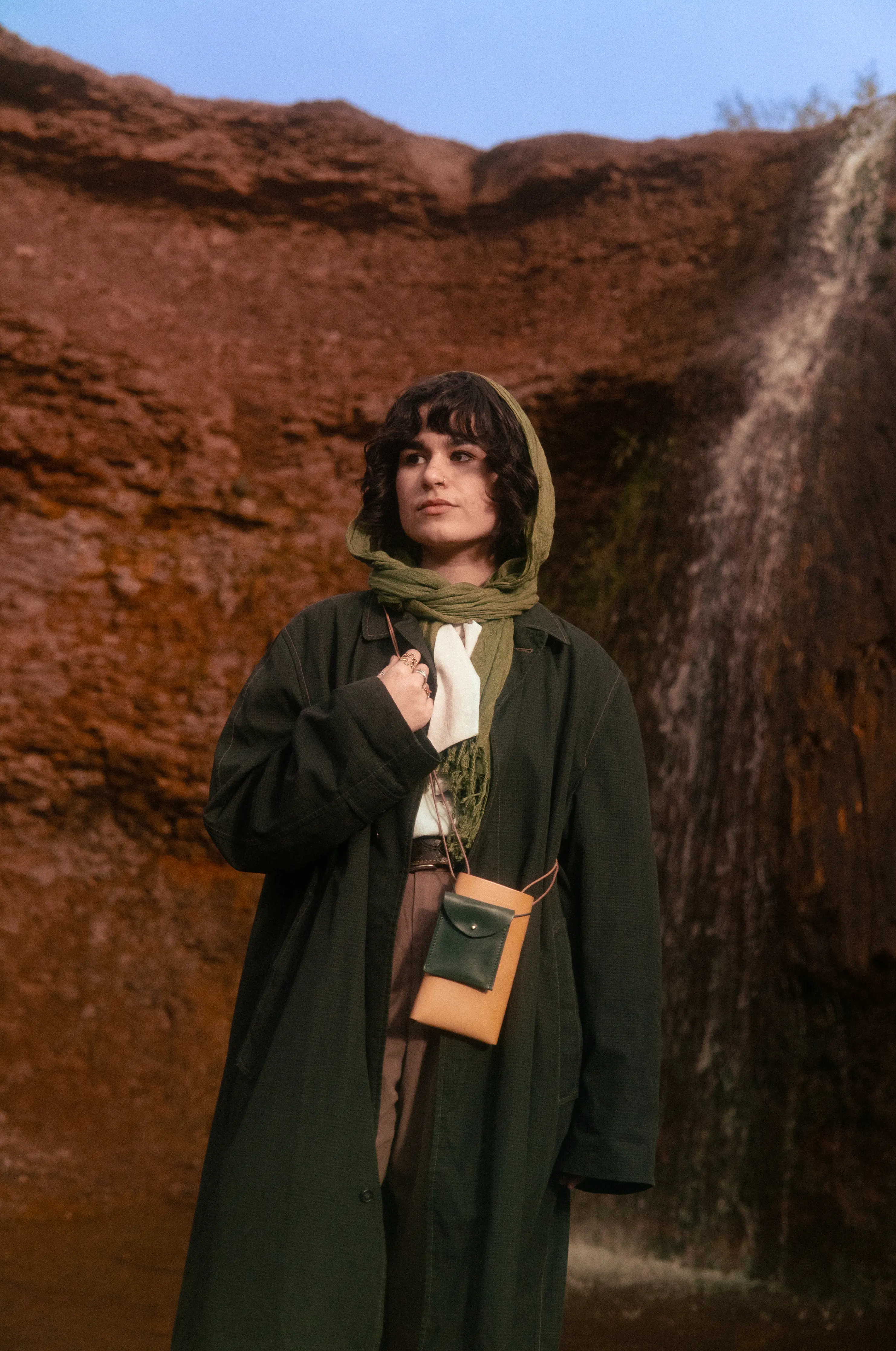 Anaïs Brancheriau wearing a green scarf, a long dark coat and a two-tone pouch, standing in front of a waterfall framed by red cliffs.