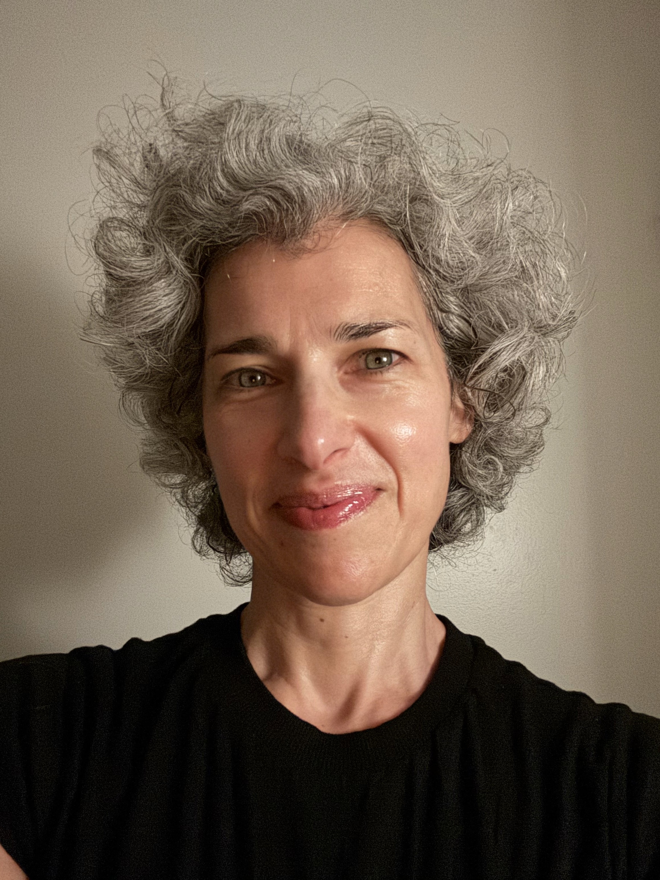 A smiley lady with grey curly hair and light green eyes wearing a black t-shirt.